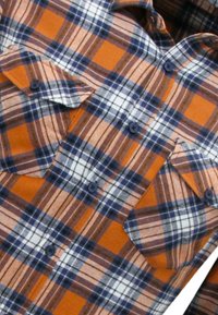 Oranje en bruin geruite flanellen shirt met blauwe accenten, twee borstzakken en knoopsluiting. Textuur lijkt zacht en de stof ziet er duurzaam uit.