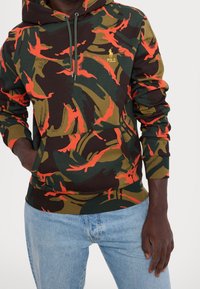 Sudadera de camuflaje verde y marrón con acentos naranjas, capucha con cordón, bolsillo canguro y detalles del logo dorado en el pecho. Corte casual.