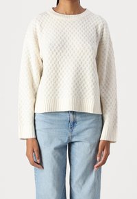 Cremfarbener gestrickter Pullover mit strukturiertem Muster, gerippten Bündchen und Saum sowie Rundhalsausschnitt, kombiniert mit hellblauen Jeans.