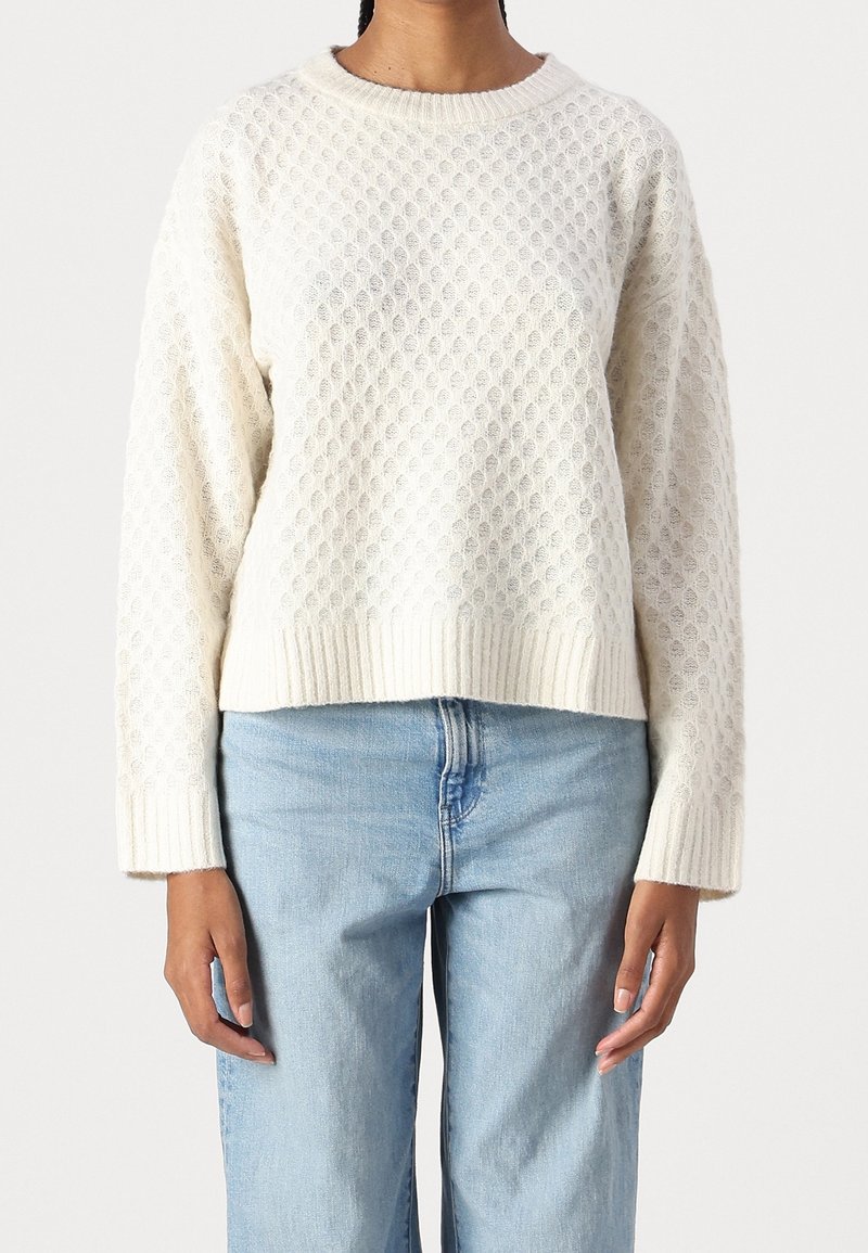 Cremfarbener gestrickter Pullover mit strukturiertem Muster, gerippten Bündchen und Saum sowie Rundhalsausschnitt, kombiniert mit hellblauen Jeans.