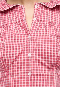 Κόκκινο και λευκό πουκάμισο με κουμπιά σε καρό σχέδιο και φαρδύ γιακά, φορεμένο μερικώς ξεκουμπωμένο, αποκαλύπτοντας την πάνω περιοχή του στήθους και του λαιμού.
