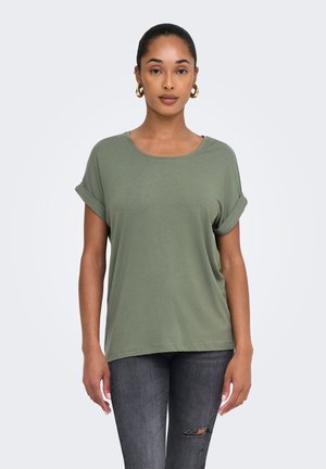 Mujer con camisa de manga corta verde oliva con puños remangados y vaqueros rotos gris oscuro, de pie frente a un fondo claro y liso.