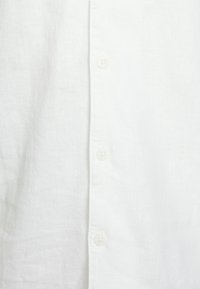 Camisa de lino blanca con un cierre de botones en la parte delantera que tiene cuatro botones a juego, una textura suave y un diseño de cuello clásico.