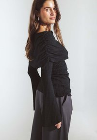 Čierne, textúrované tričko s dlhým rukávom, so zloženými detailmi a rozšírenými rukávmi, skombinované s voľne strihanými sivými nohavicami.