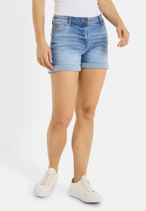 Mujer con shorts vaqueros azules claros remangados, camiseta sin mangas blanca y zapatillas blancas, de pie frente a un fondo liso.
