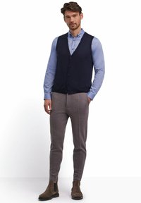Gilet lavorato a maglia blu navy sopra una camicia azzurro chiaro con bottoni, abbinato a pantaloni grigi a sigaretta e stivali alla caviglia in suede marrone.