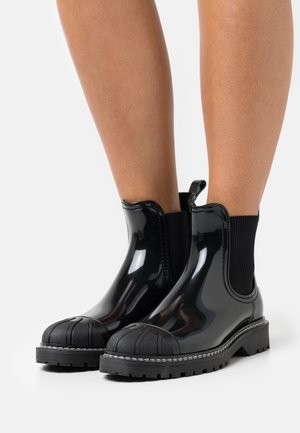 Zwarte glanzende enkellaarsjes van rubber met een gestructureerde neuskap, elastische zijkanten en een robuuste zool, met witte stikselaccenten.