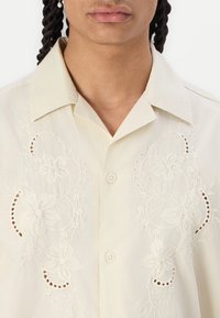 Camisa de botón en crema claro con bordado floral y detalles de ojales en la parte delantera. Tejido suave, cuello clásico y corte recto.