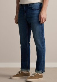 Jeans de mezclilla azul para hombre con un corte recto, que presentan un diseño clásico de cinco bolsillos y dobladillos, combinados con zapatillas beige.
