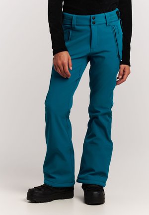 Personne portant un pantalon d'extérieur bleu sarcelle avec deux poches zippées et des chaussures de randonnée noires sur un fond clair uni.