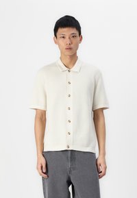 Polo à manches courtes de couleur crème en tricot texturé avec un col et une fermeture à boutons beige ; associé à un pantalon gris.