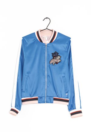 Niebieska satynowa kurtka typu bomber z białymi paskami na rękawach, kwiatową haftem na piersi oraz prążkowanym kołnierzem, mankietami i dolnym obszyciem w paski, wisząca na czarnym wieszaku.