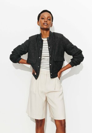 Femme debout, mains sur les hanches, portant une veste bomber noire texturée, une chemise rayée et un short beige arrivant aux genoux, sur fond blanc.