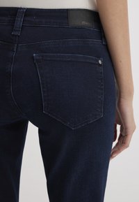 Dark blue denim jeans featuring a classic five-pocket design, textured fabric, and a leather logo patch on the back waistband.