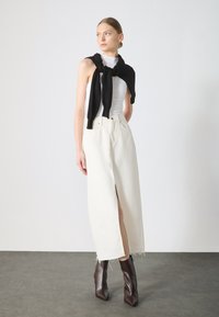 White denim skirt with a front slit, paired with a fitted, ribbed white top. Black sweater tied at the neck. Brown square-toed heeled boots.