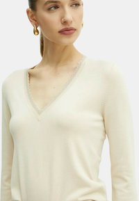 Pullover beige chiaro con maniche lunghe, scollatura a V e bordo ondulato. Texture morbida e design aderente, adatto per essere indossato a strati.