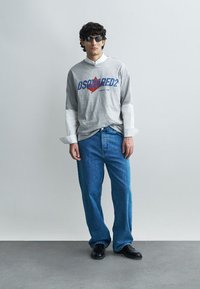 Pelēks garā piedurkņu T-krekls, kārtots virs baltas krekla, kopā ar zilām džinsa biksēm un melniem apaviem. T-kreklam ir uzraksts "DSQUARED2."