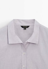 Chemise gris clair à boutons avec un col classique, de fines rayures verticales et une étiquette noire visible à l'intérieur du col.