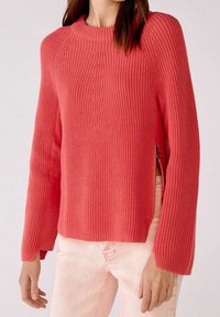 Pull en tricot corail avec texture côtelée, col rond et fentes sur les côtés ; associé à un jean rose clair. Détail de couture visible sur le devant.