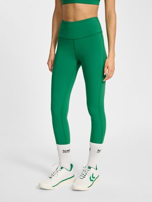 Vrouw draagt groene leggings met hoge taille, witte sportsokken met zwarte letters en witte sneakers met groene accenten, staand tegen een vlakke achtergrond.