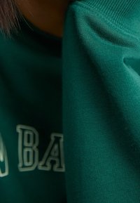 Felpa verde dal tessuto morbido, con maniche arrotolate e lettere bianche. Vista ravvicinata che mette in evidenza i dettagli delle cuciture.