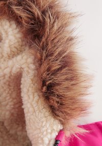 Pruun kunstkarusnahast ääris ümbritseb kreemjat fliisvoodrit, mis on kinnitatud erkroosa välisriide külge. On nähtav must metalldetail.