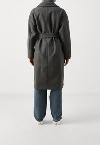 Grå ullkappa med bälte, avslappnad passform, långa ärmar och slagkrage. Buren över blå vida jeans och vita sneakers.