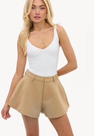 Mujer rubia con una camiseta blanca sin mangas con escote en V y pantalones cortos beige de talle alto con bolsillos, de pie contra un fondo liso.