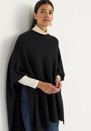 Frau steht vor einer weißen Wand, trägt einen schwarzen Poncho, ein weißes langärmeliges Oberteil und blaue Jeans, die Hände sanft verschränkt.