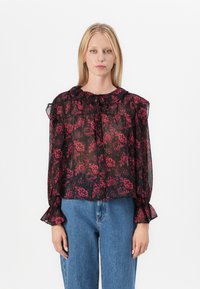 Blusa nera trasparente con motivo floreale rosso, scollatura arricciata e maniche a campana, abbinata a jeans a vita alta blu.
