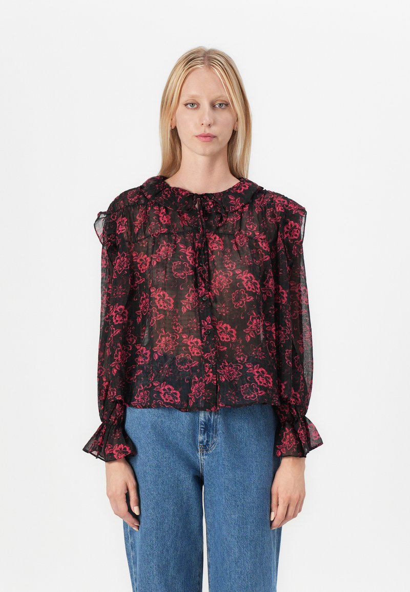 Blusa nera trasparente con motivo floreale rosso, scollatura arricciata e maniche a campana, abbinata a jeans a vita alta blu.