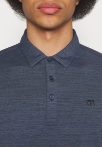 Mørkeblå polo-skjorte med strukturert mønster, med spredd krage, tre knapper og et lite brodert logo på venstre bryst.