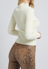 Haut ivoire à manches longues côtelé avec col montant, associé à un pantalon marron ajusté présentant un motif subtil de peau de serpent et une étiquette en cuir sur la poche.