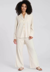 Lichtbeige, overhemd met lange mouwen en knopen, voorzien van een borstzak, gecombineerd met bijpassende wijde broek. De knopen hebben een gouden kleur.