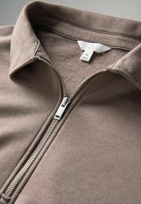 Taupe hnedá bunda na zips so strieborným kovovým zipsom a prehnutým golierom, ktorý ukazuje bielu etiketu s veľkosťou M vo vnútri výstrihu.
