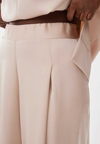 Pantalons beiges en tissu lisse, avec des plis sur les côtés et une large ceinture élastique pour le confort et un ajustement soigné.