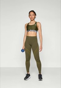 Fiatal nő olajzöld sportmelltartóban és leggingsben, kék súlyzóval a kezében, egy egyszínű világos háttér előtt áll.