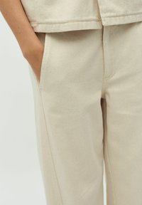 Pantalons en coton beige à coupe droite, avec des poches latérales et une finition douce et texturée. La taille est bien ajustée.