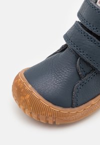 Zils ādas apavs ar teksturētu virsmu, kurā ir divi velcro siksniņas un gumijas zole ar ribinātu detaļu.