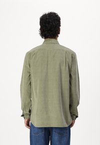 Licht olijfgroene blouse met lange mouwen, een kraag en achterplooien, gemaakt van een zachte stof. Gecombineerd met blauwe spijkerbroek.