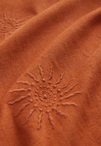 Rust-colored fabric featuring subtle sun embroidery with circular center and radiating lines, showcasing a textured, natural material.