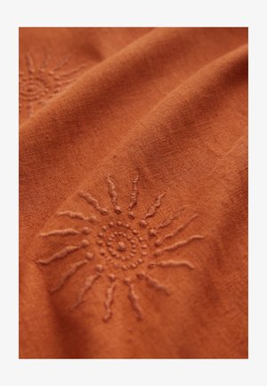 Rust-colored fabric featuring subtle sun embroidery with circular center and radiating lines, showcasing a textured, natural material.