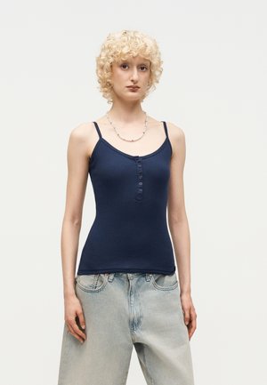 Person med krøllete blondt hår iført en marineblå knappet tanktop og lyse, vide jeans, står foran en ensfarget bakgrunn.