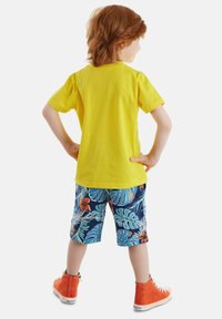 Camiseta amarilla de manga corta, pantalones cortos azules con un estampado de hojas y flores tropicales, y zapatillas rojas de tobillo con suelas blancas.