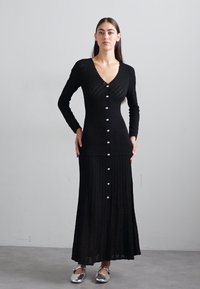 Zwarte lange mouwen gebreide jurk met een V-hals en diamantstructuur, met knopen langs de voorkant en een geplooide rok. Zilveren schoenen.