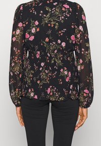 Blusa nera floreale con maniche lunghe, dotata di vita arricciata e vivaci motivi di fiori rosa e verdi su un tessuto semitrasparente.