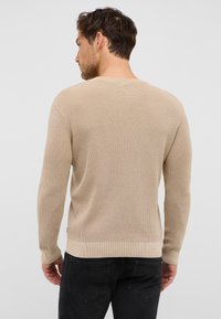 Beżowy sweter dziergany z prążkowaną fakturą, okrągłym dekoltem i długimi rękawami. Posiada dopasowany pas i mankiety. Nosi się go w połączeniu z ciemnymi dżinsami.