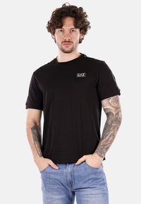 Camiseta de algodón negra con cuello redondo, mangas cortas y una pequeña etiqueta de logo. Combinada con unos jeans de mezclilla azul claros.