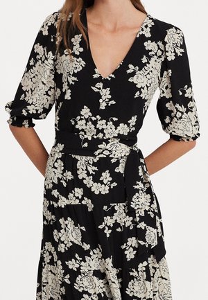 Vrouw draagt een zwarte wikkeljurk met witte bloemenprint, V-hals, pofmouwen en een gestrikte tailleband.