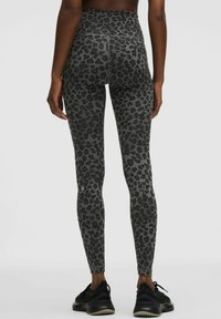 Mujer vista desde atrás vistiendo leggings de talle alto con estampado de leopardo gris y zapatos deportivos negros sobre un fondo gris liso.